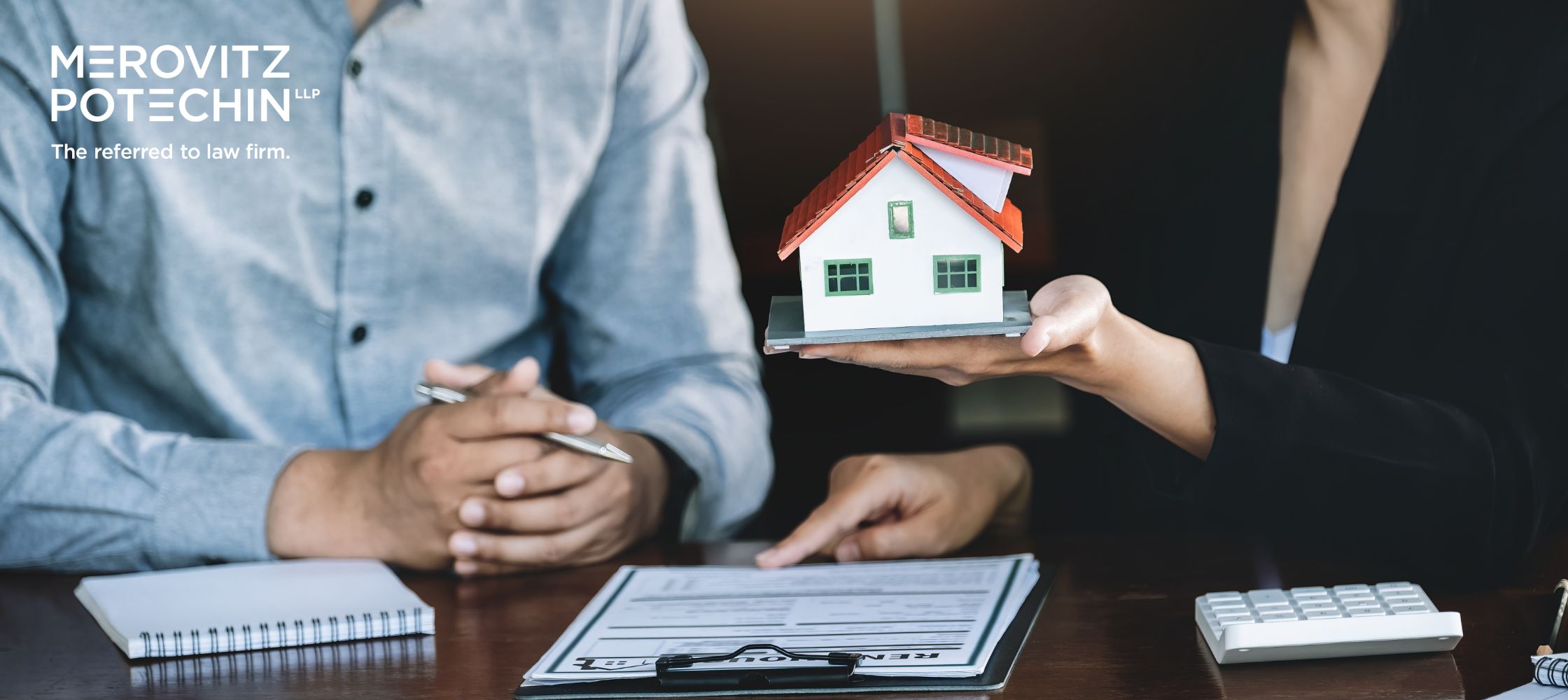 A lawyer and client reviewing real estate documents related to Agreements of Purchase and Sale, with a model house placed on the table to represent a property transaction.