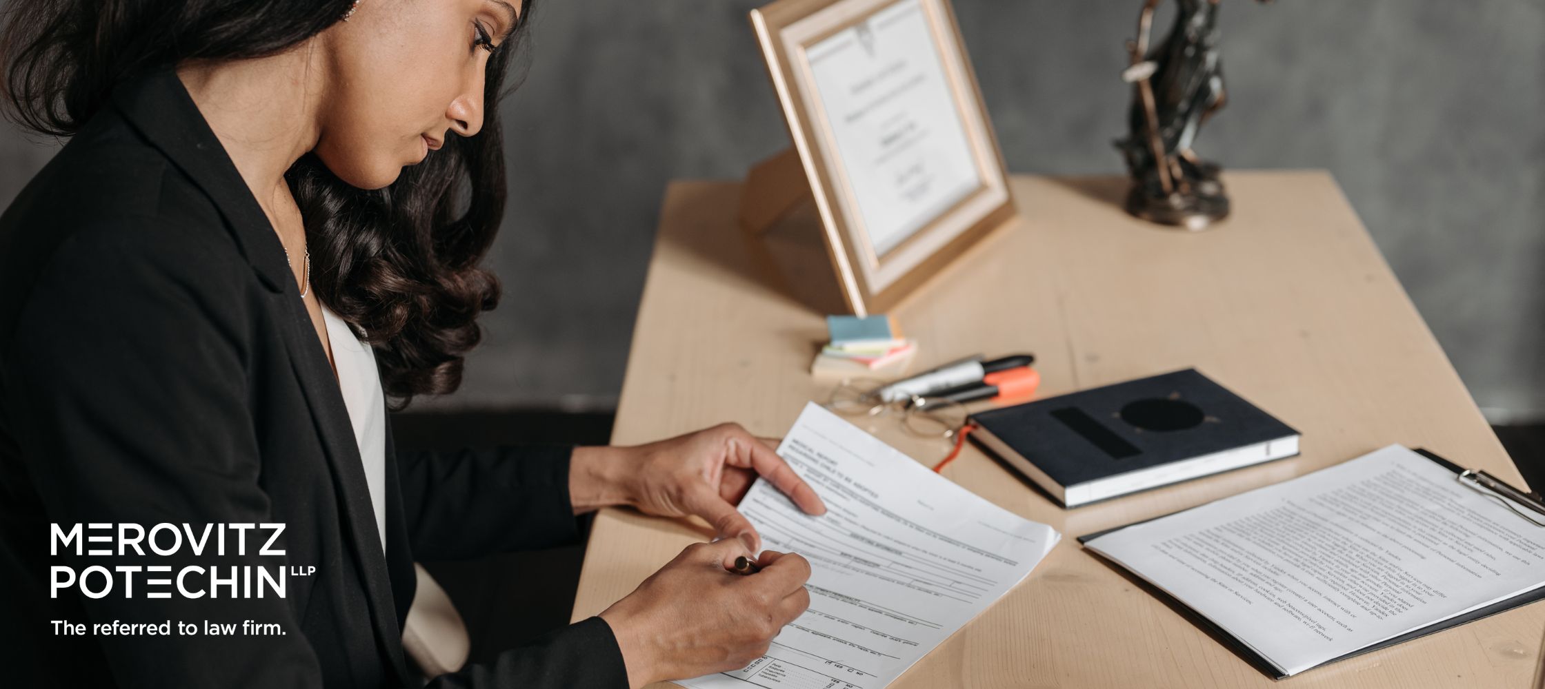 A female articling student reviewing and writing on legal documents at a wooden desk with a scales of justice statue and the Merovitz Potechin LLP logo.